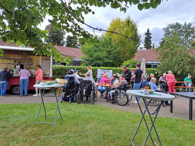 Zu sehen ist ein Imbisswagen. Daneben stehen mehrere Tische, an denen Bewohnende und ihre Gäste feiern.