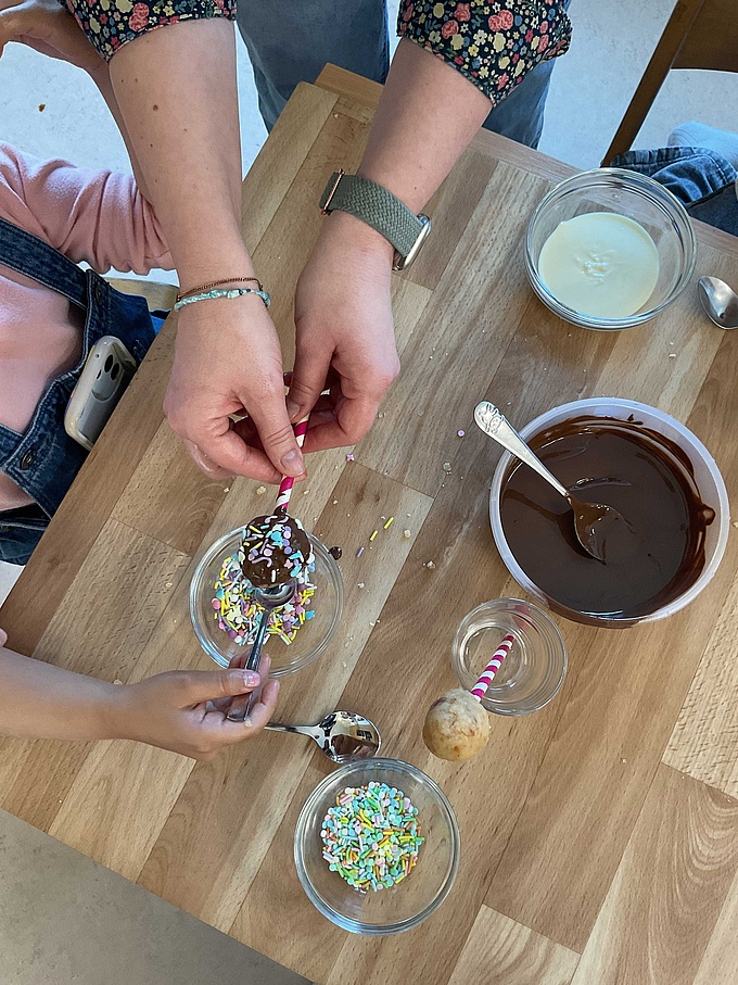 Eine Erzieherin und ein Kind machen bunte Cake Pops mit Schokolade und Streuseln.