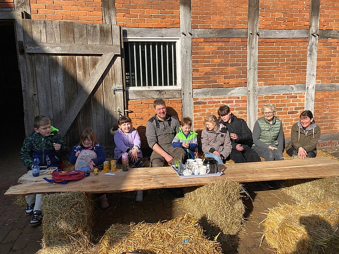 Die Schüler*innen, die Hofbesitzer und die Naturschutzführerin Christiane Achelwilm sitzen auf einer selbstgebauten Bank aus Strohballen und Brettern und frühstücken.