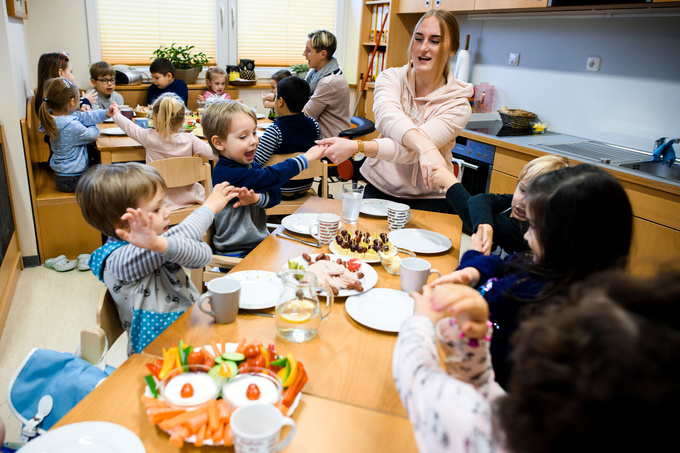 Essenstraditionen: Eine Erziherin und 4 Kinder sitzen am Esstisch und halten sich mit überkreuzten Armen an den Händen.