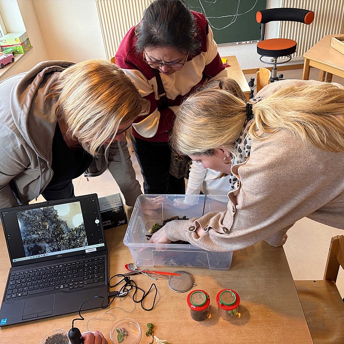 Drei Frauen beobachten eine Bodenprobe. Vor ihnen steht ein Laptop, der die Bodenprobe optisch vergrößert. 