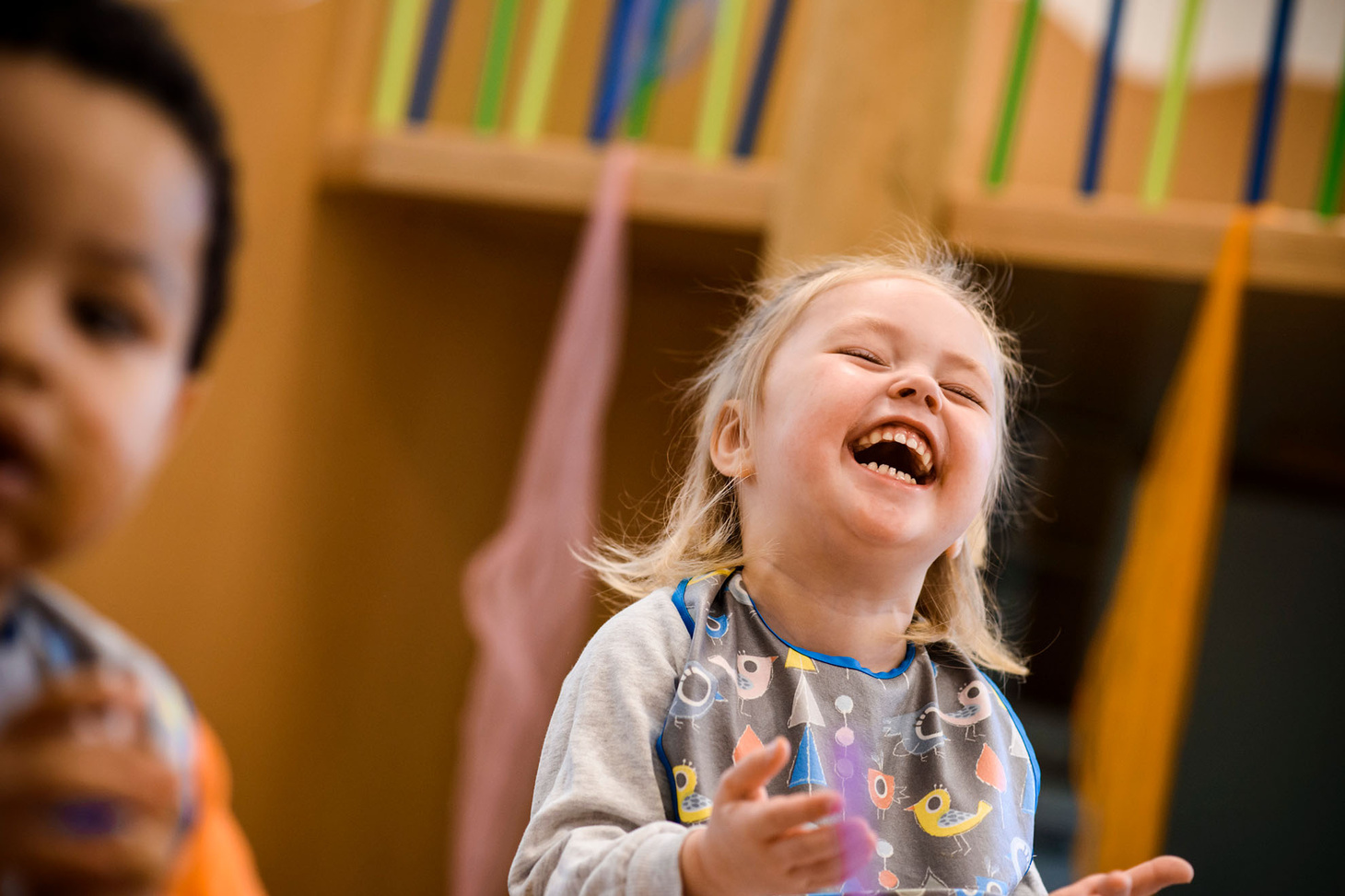 Ein weißes Kind mit blonden Haaren lacht herzhaft. Im Vordergund ist verschwommen ein Schwarzes Kind mit kurzem Haar zu sehen.