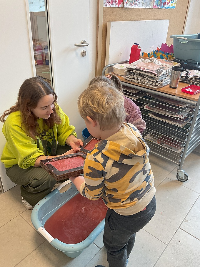 Zwei Kinder und Isabella Schablinski betrachten ein spezielles Sieb, mit dem neue Papierbögen geschöpft werden. 