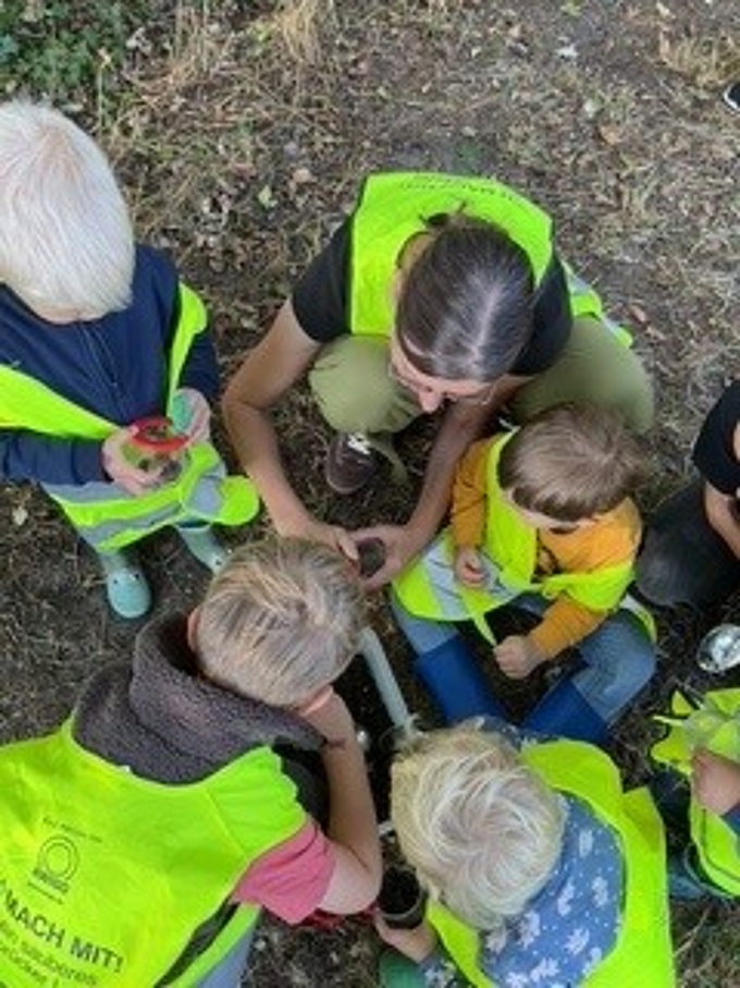 Mehrere Kinder und Isabella von der Pädagogischen Abfallberatur des Landkreises hocken auf dem Boden und nehmen Erde unter die Lupe. 